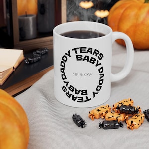 May include: White ceramic mug with black text that reads "DADDY TEARS BABY" in a circular pattern, with "SIP SLOW" in the center. The mug is filled with a dark liquid. Halloween-themed candies are scattered around the mug.
