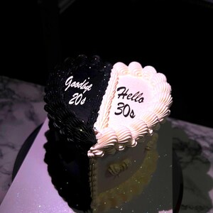 May include: A heart-shaped cake, half black and half white, with the words "Goodbye 20s" and "Hello 30s" in white script. The cake has a decorative border and sits on a silver surface.