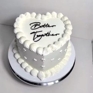 May include: A white heart-shaped cake with white frosting and a black inscription that reads "Better Together".