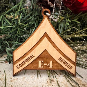 May include: Wooden Christmas ornament shaped like a military rank insignia, with the words "Corporal E-4 Dawson" engraved on it.