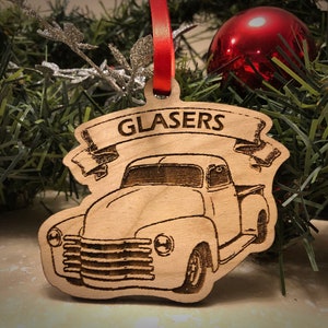 May include: A wooden Christmas ornament shaped like a vintage pickup truck with the name "GLASERS" on a banner above it. The ornament is painted in a natural wood color.