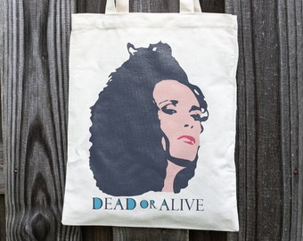 May include: A white canvas tote bag with a black and white illustration of a woman with dark curly hair. The text "DEAD OR ALIVE" is printed in blue below the illustration.