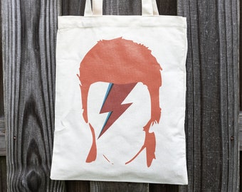 May include: A white canvas tote bag with a red and blue lightning bolt design. The design is a silhouette of David Bowie's face.