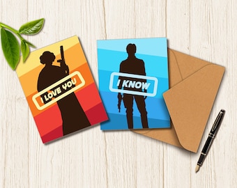 May include: Two greeting cards with silhouettes of characters, one with the text "I LOVE YOU" and the other with "I KNOW". The cards are on a wooden surface with a brown envelope and a black pen.