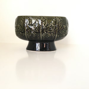 Puede incluir: Cuenco de cerámica verde oscuro con base de pedestal. El cuenco presenta un patrón de hojas repetido alrededor del borde. El acabado brillante refleja la luz, resaltando el diseño detallado. Una pieza decorativa para la decoración del hogar.