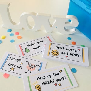 May include: A selection of motivational lunchbox notes with phrases such as "Enjoy your lunch" and "Don't worry, be HAPPY!" are displayed. A white "LOVE" sign and blue confetti are also present. A blue lunchbox is in the background. The notes are on a white surface.