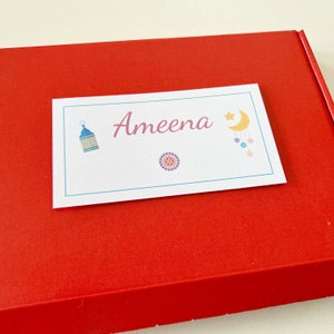 May include: A white card with the name "Ameena" in pink script, featuring a blue lantern, a colourful mandala, and a crescent moon and star design. The card rests on a red box.