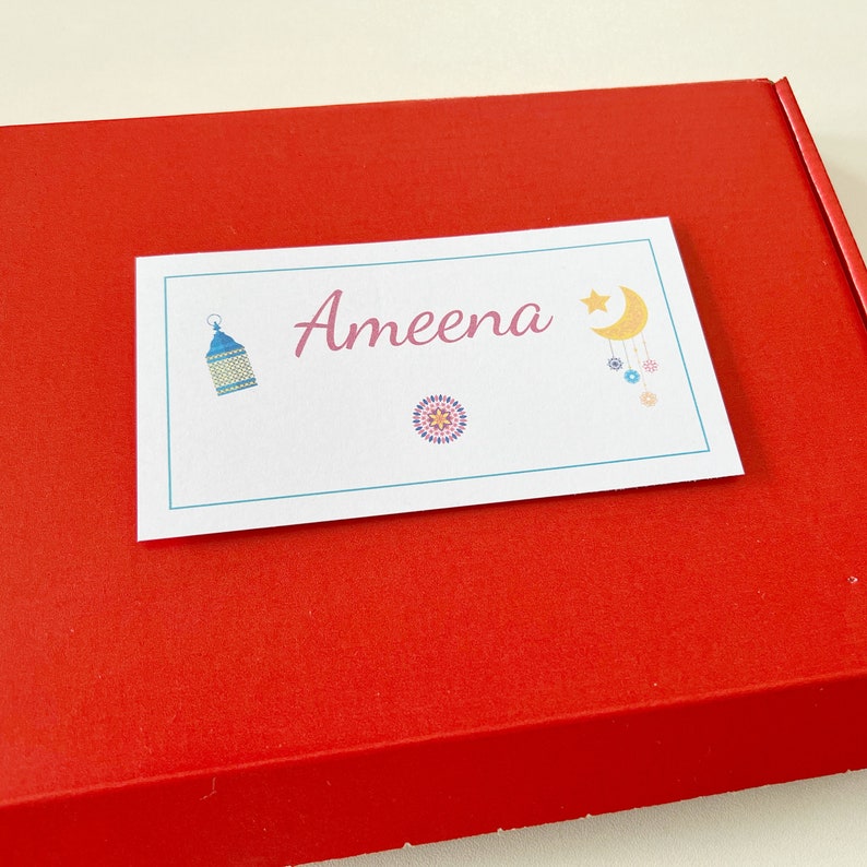 May include: A white card with the name "Ameena" in pink script, featuring a blue lantern, a colourful mandala, and a crescent moon and star design. The card rests on a red box.