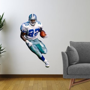May include: A wall decal of a football player in a blue and white uniform, holding a football. The player is in mid-run, with a silver helmet. A green plant in a white pot and a gray couch are also in the image.