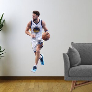May include: A basketball player wearing a white and blue jersey with the Golden State Warriors logo dribbling a basketball. The player is in mid-air, with one leg extended and the other bent. The player is wearing blue and white trainers. The player is in front of a white wall with a grey sofa in the background.