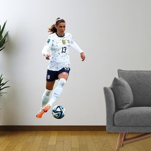 May include: A female football player wearing a white and black jersey with the number 13 on it, white socks, and black shorts, is kicking a football. The player is in mid-air and the ball is in front of her. The background is a white wall.