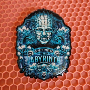 May include: A blue and black enamel pin featuring a character with a face covered in pins and the text "Welcome to the Labyrinth".