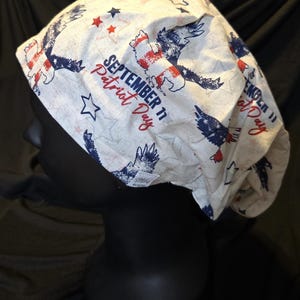 May include: A white surgical scrub cap featuring patriotic imagery. The cap is adorned with red, white, and blue eagles, stars, and the text "SEPTEMBER 11 Patriot Day". The design is printed on a light-colored fabric.
