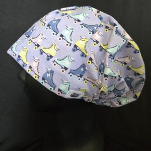 May include: A light purple surgical scrub cap featuring a repeating pattern of colorful roller skates in shades of blue, yellow, and pink. The cap is made of a soft fabric and has a small white tag with a logo.