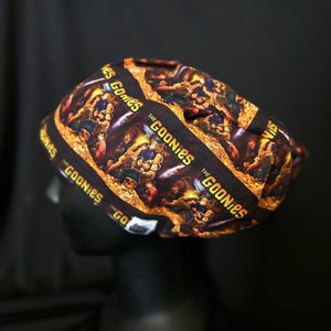 May include: A black cap featuring a repeating pattern with the text "THE GOONIES" in yellow and a movie scene graphic. The cap is displayed on a black mannequin head against a dark background.