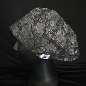 May include: A black and white patterned scrub cap featuring a forest tree design. The cap is displayed on a black mannequin head. A small white tag with black text is attached to the cap.
