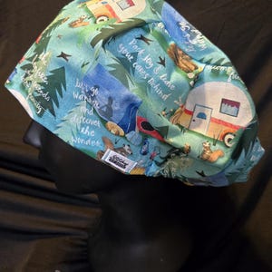 May include: A teal camping-themed scrub cap featuring illustrations of campers, trees, and squirrels. The cap has text that reads "Pack joy & leave your cakes behind" and "Let's go wander and discover the wonder."