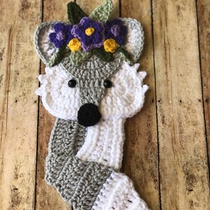 May include: A crocheted fox with a white tail and gray body. The fox is wearing a flower crown with purple and yellow flowers.