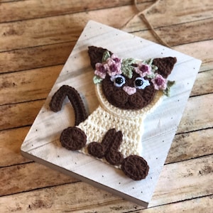 May include: A crocheted cat with brown and white yarn, wearing a flower crown with pink and green flowers, is attached to a white wooden plaque.