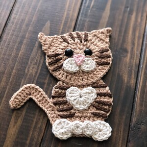 May include: A crocheted brown and white cat with black eyes and a pink nose. The cat is holding a white heart in its paws and has white feet.