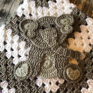 May include: A crocheted baby blanket with a brown elephant applique. The blanket is made with white and brown yarn and has a granny square pattern.