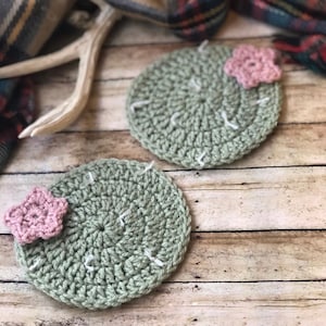May include: Two green crocheted cactus coasters with pink flower accents. The coasters are on a wooden surface.