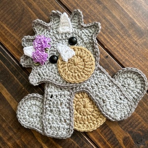 May include: A crocheted grey triceratops with brown eyes and a purple flower on its head. The triceratops is sitting on a wooden surface.