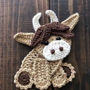 May include: A handmade crochet cow applique in shades of tan, brown, and cream. The cow has black eyes, a white muzzle, and cream-colored horns. The applique is designed with detailed crochet stitches and is set against a dark wood background.