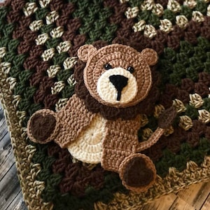 May include: A handmade crochet baby blanket featuring a lion design. The blanket is a mix of brown, green, and cream colors. The lion has a brown mane, ears, and paws, with a cream-colored face and a black nose. The blanket is square-shaped.