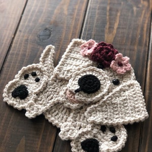 May include: A crocheted dog with a flower crown. The dog is beige with black eyes and nose. The flower crown is made of burgundy and pink flowers.