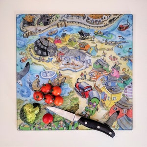 May include: A colourful glass chopping board with a whimsical illustration of a seaside town. The board features a variety of houses, boats, and people, all drawn in a cartoon style. There are also some red tomatoes and a green broccoli floret on the board, along with a silver knife.