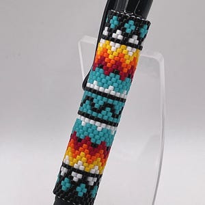 May include: A black ink pen featuring a barrel covered in a woven bead pattern. The design consists of repeating geometric shapes in vibrant colors including turquoise, white, orange, yellow, and red, all outlined with small black beads.