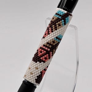 May include: A black ink pen featuring a cylindrical barrel covered in a woven bead pattern. The beads are arranged in a geometric design using a color palette of white, dark brown, light pink, and bright turquoise, creating a repeating motif.