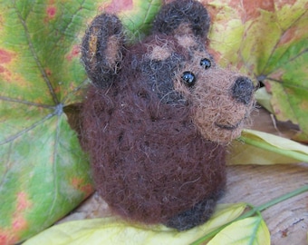 Pode incluir: Um pequeno urso marrom feito de feltro com olhos pretos. O urso está sentado em uma cama de folhas de outono.