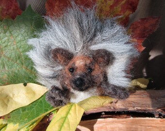 May include: A close-up of a needle felted hedgehog with a white and gray spiky coat. The hedgehog is sitting on a bed of brown and yellow leaves.
