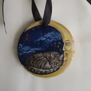 May include: A wooden ornament painted with a crescent moon, a sleeping cat, and a face. The moon is yellow, the cat is brown and gray, and the face is pale with a yellow halo. The ornament is tied with a black ribbon.