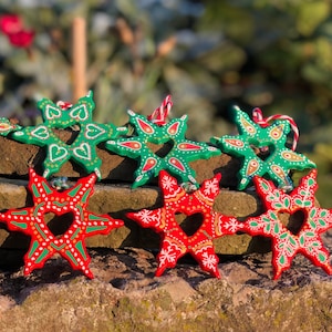 May include: Eight hand-painted wooden star ornaments, four green and four red, with intricate designs and a heart-shaped cutout in the center. The stars are arranged on a stack of brown stones.