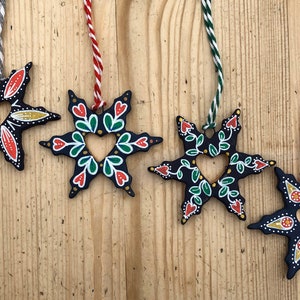 May include: Five black wooden star ornaments with a heart-shaped cutout in the center. Each star is painted with different colors and patterns, including red, green, yellow, and white.