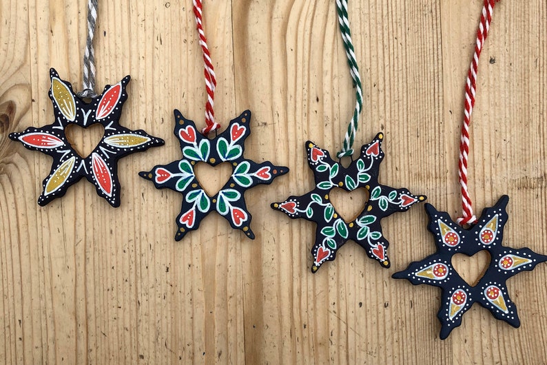 May include: Five black wooden star ornaments with a heart-shaped cutout in the center. Each star is painted with different colors and patterns, including red, green, yellow, and white.
