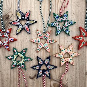 May include: Nine hand-painted wooden star ornaments with a heart-shaped cutout in the center. Each star is painted in a different color combination, including red, green, blue, yellow, and black. The stars are hanging from twine.