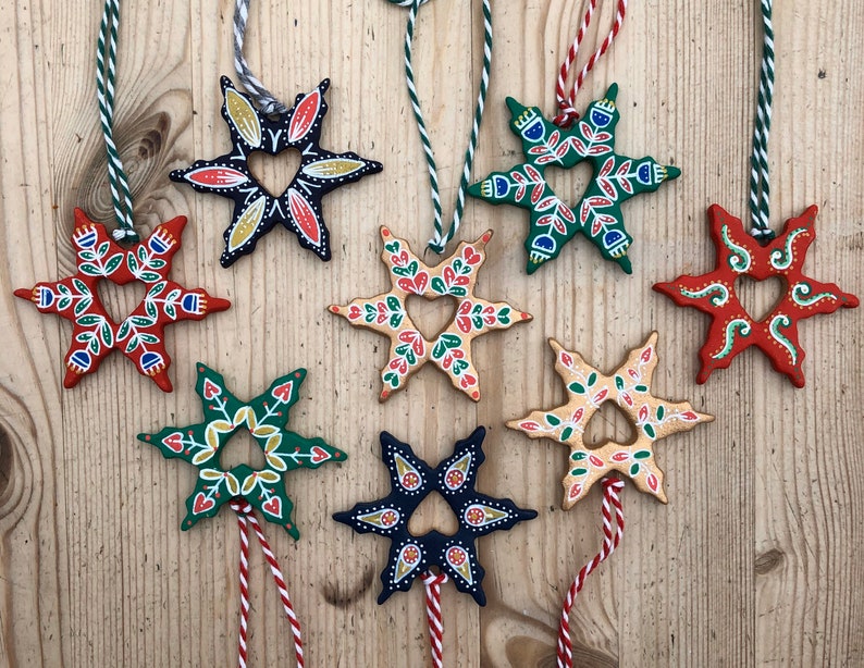 May include: Nine hand-painted wooden star ornaments with a heart-shaped cutout in the center. Each star is painted in a different color combination, including red, green, blue, yellow, and black. The stars are hanging from twine.