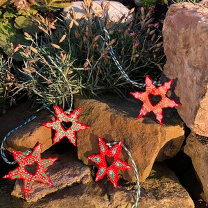 May include: Four red star-shaped ornaments with green and white painted details. Each star has a heart-shaped cutout in the center. The stars are hanging from twine.