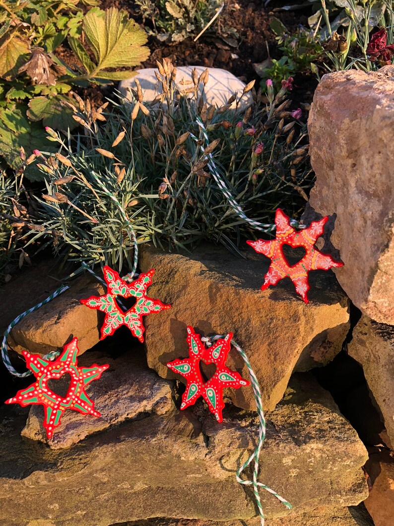 May include: Four red star-shaped ornaments with green and white painted details. Each star has a heart-shaped cutout in the center. The stars are hanging from twine.