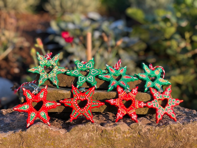 May include: Eight hand-painted wooden star ornaments, four green and four red, with intricate designs and a heart-shaped cutout in the center. The stars are arranged on a stack of brown stones.