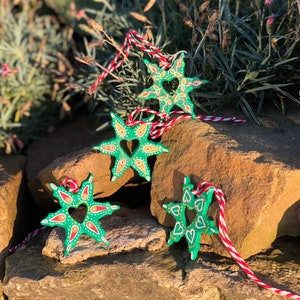 May include: Four green star-shaped ornaments with a heart design and white and orange accents. The ornaments are hanging from red and white string.