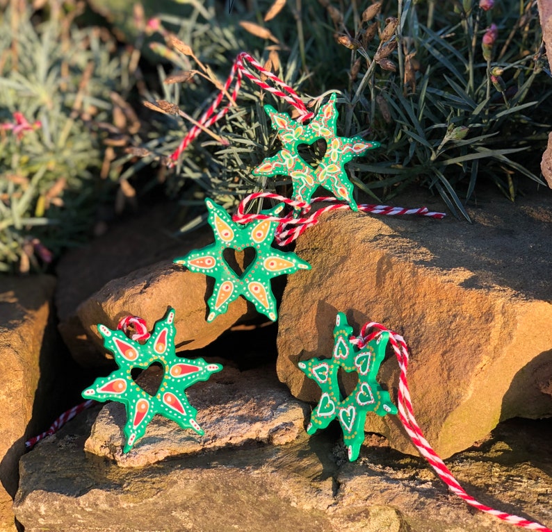 May include: Four green star-shaped ornaments with a heart design and white and orange accents. The ornaments are hanging from red and white string.