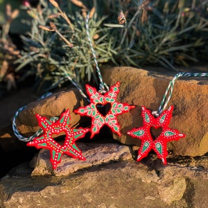 May include: Three red wooden snowflake ornaments with green and white painted details. Each snowflake has a heart-shaped cutout in the center. The snowflakes are hanging from white and green twine.