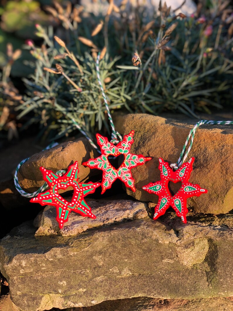 May include: Three red wooden snowflake ornaments with green and white painted details. Each snowflake has a heart-shaped cutout in the center. The snowflakes are hanging from white and green twine.