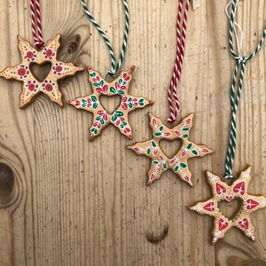 May include: Five gold star-shaped ornaments with red and green painted details. Each ornament has a heart-shaped cutout in the center. The ornaments are hanging from strings.