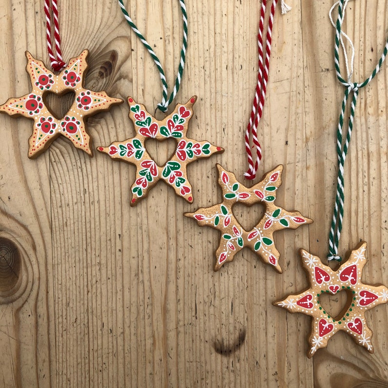 May include: Five gold star-shaped ornaments with red and green painted details. Each ornament has a heart-shaped cutout in the center. The ornaments are hanging from strings.